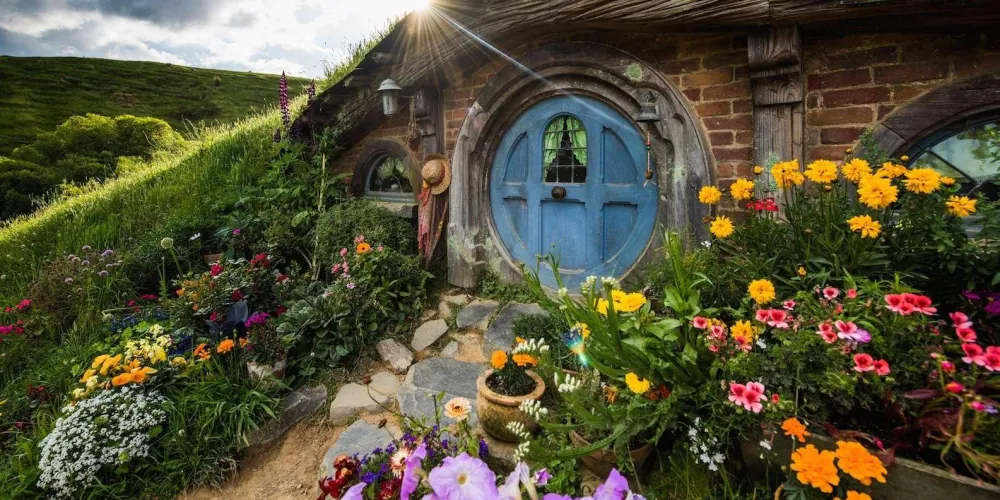 A round blue hobbit-hole door built into a grassy hillside, surrounded by a tended cottage garden, on the Hobbiton Movie Set